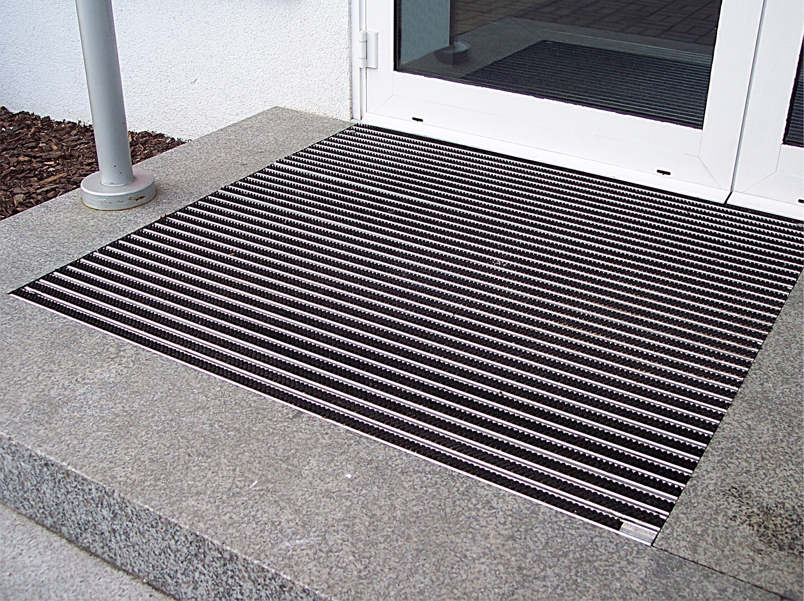 Black Venture Brush entrance grid installed in a stone-paved floor recess outside an entrance